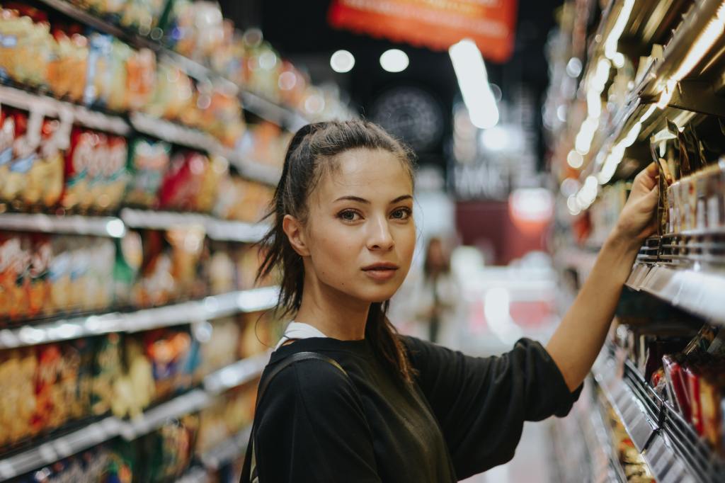 Woman with brown hair in a ponytail reaching up to supermarket shelf and looking to her right, at the camera.