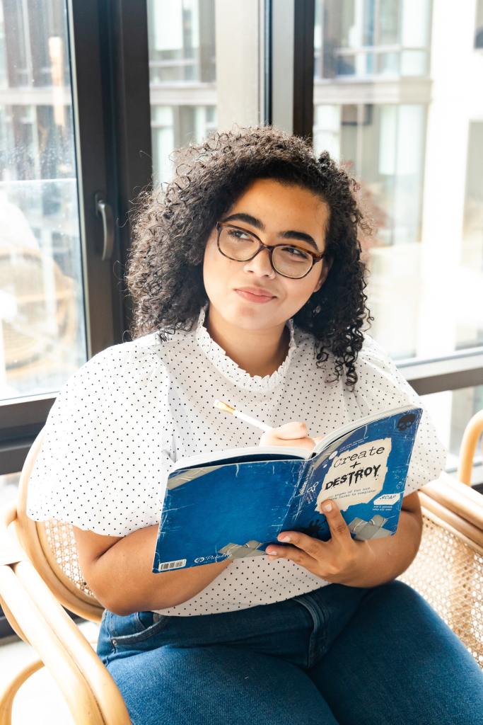 Seated woman with curly hair and glasses facing the camera and looking to the side with a thoughtful smile on her face, and pen poised to write in her notebook, called 'Create + Destroy'