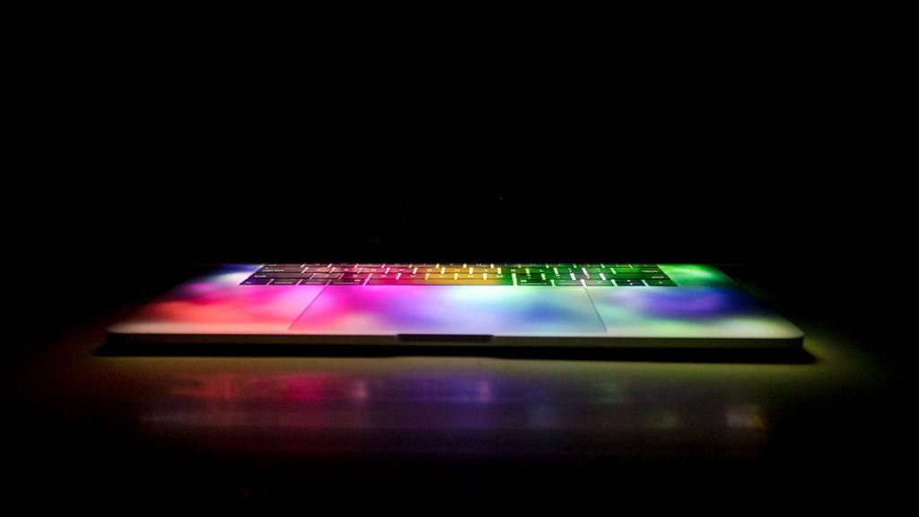 Photo of a laptop keyboard on black background with rainbow lights projected onto it.